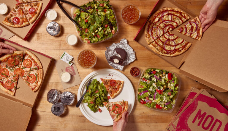 A MOD Pizza catering bundle lays spread out across a table, with numerous No Name Cake desserts, different signature pizzas, and salads to go along with it.
