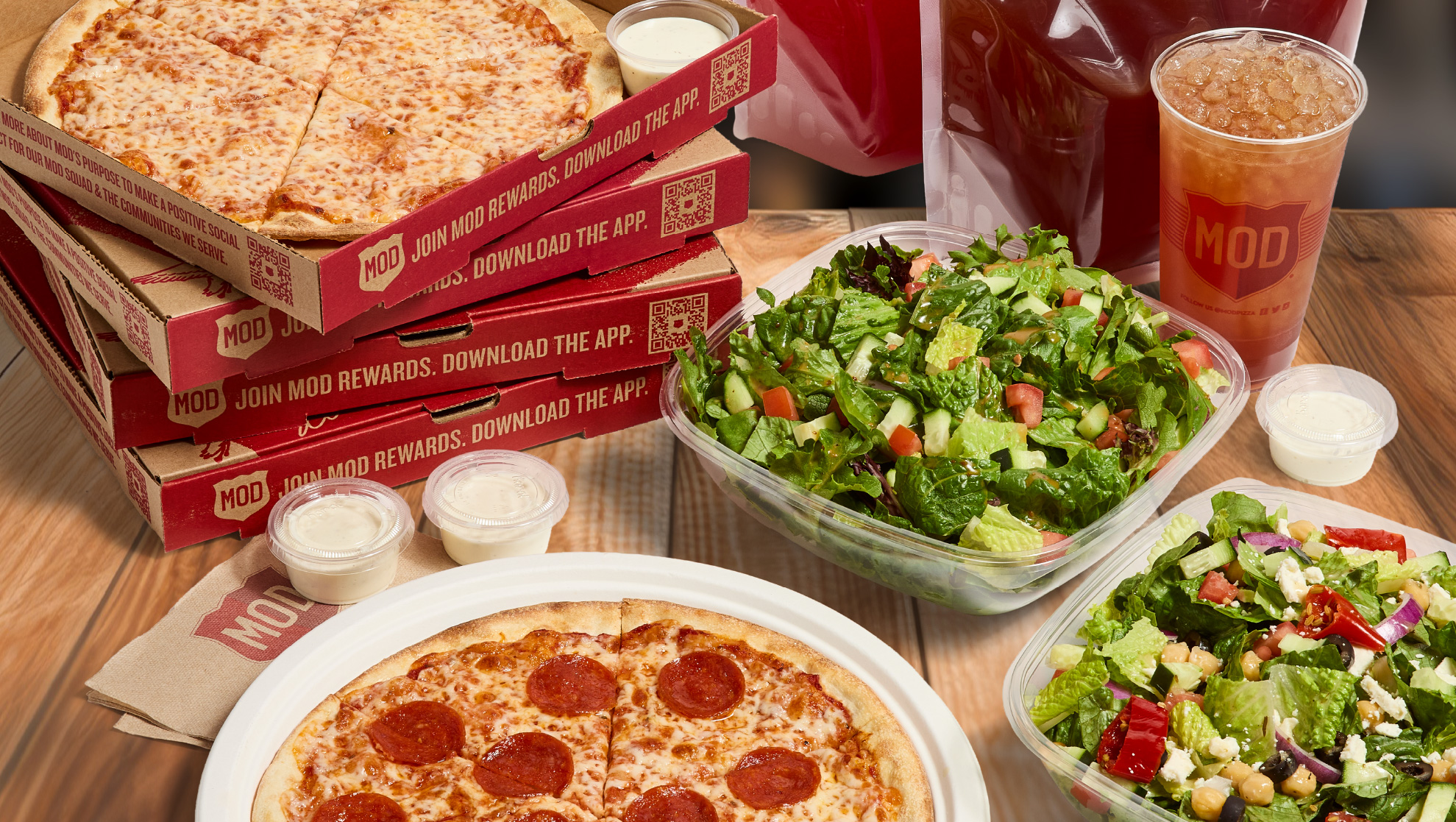 A MOD Pizza Signature Catering Bundle with open pizzas, salads, and drinks, all sitting on a wooden table.